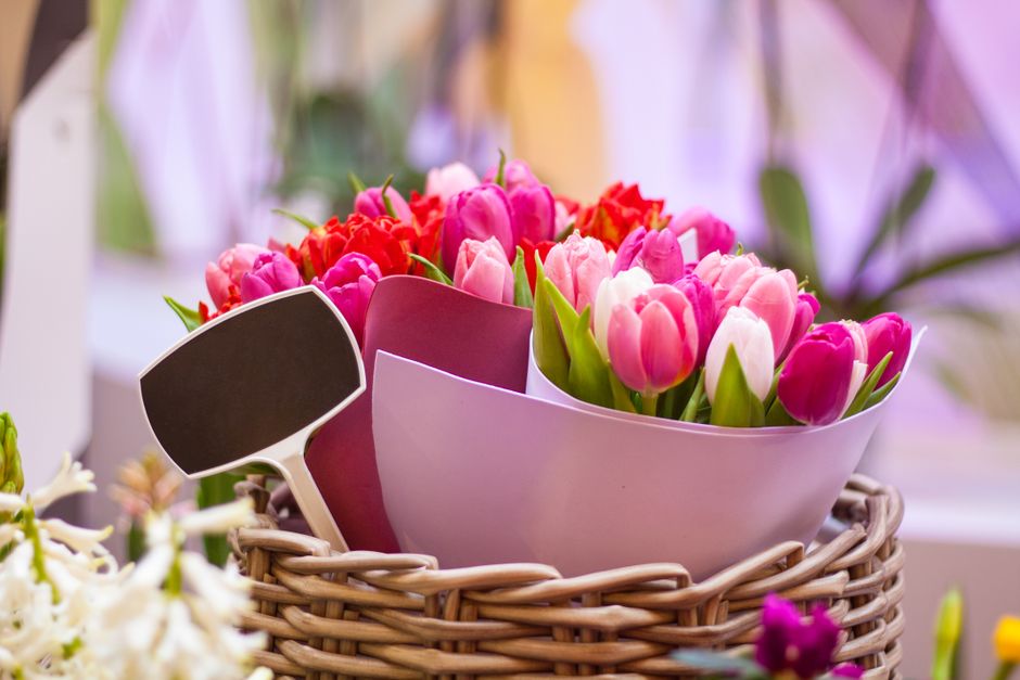 A floral arrangement featuring an assortment of fresh pink and white tulips along with the vibrant red blossoms of freesias, all arranged in a white ceramic bowl. The bouquet is nestled inside a woven