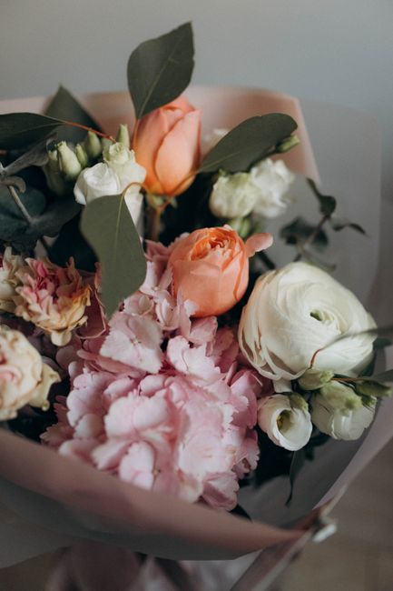 A close-up view of a floral bouquet featuring soft peach, white, and blush pink flowers, including roses and lisianthus, complemented by green eucalyptus leaves. The flowers are arranged in a stylish,