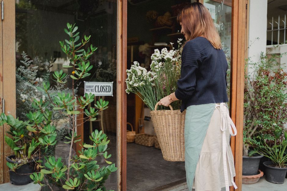 A woman with shoulder-length auburn hair, dressed in a dark blue sweater and a light-colored apron, is standing outside a flower shop holding a large wicker basket filled with fresh white and light pu