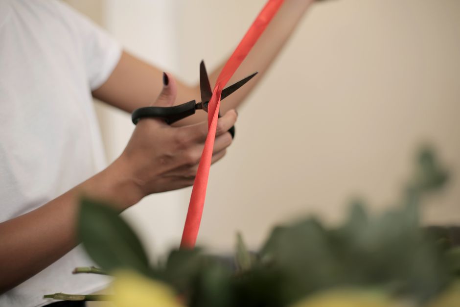 Close-up of a person’s hands holding a pair of black scissors, cutting a length of coral-colored wrapping paper around a floral arrangement. The arrangement features fresh flowers with visible petal