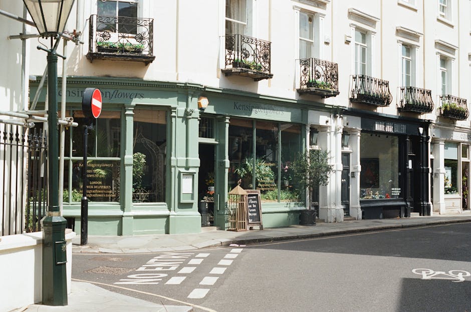 The image depicts the exterior of a florist shop located on a busy street, with the storefront painted in a muted pastel green and large glass windows showcasing various flower arrangements inside. Th