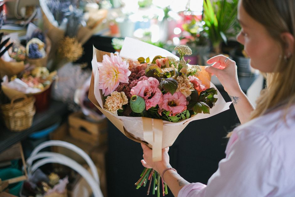 A woman with long hair is holding a vibrant bouquet of fresh flowers, including pink dahlias, roses, and various greenery, wrapped in light tan and white floral paper with a beige ribbon. She is stand