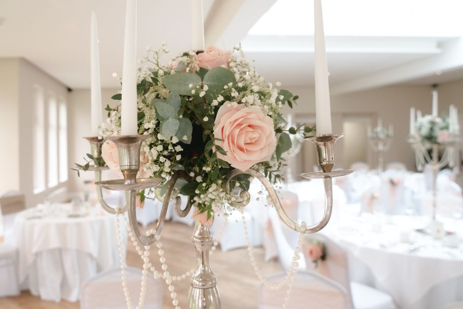 A floral arrangement featuring a bouquet of soft pink roses, white baby's breath, and eucalyptus leaves, placed on a silver candelabrum decorated with hanging pearl strands. The bouquet appears fresh 