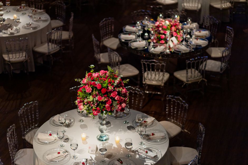 A round dining table set for a formal event, decorated with a tall, vibrant floral centerpiece composed of pink roses, green foliage, and smaller filler flowers in a clear glass vase. The table is dre